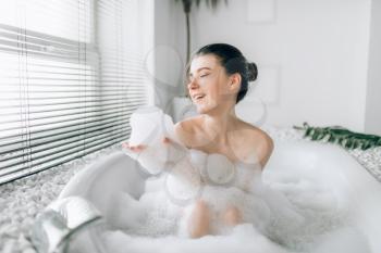 Smiling woman sitting in bathtube and plays with foam. Luxury bathroom with window and palm branch decor