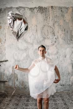 Naked freak man wrapped in packaging film holds a shiny balloon in his hands, grunge room interior. Crazy male person in abandoned house