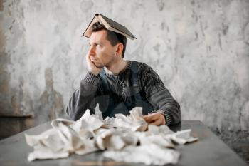 Freak man with book on his head against pile of torn sheets. Alone guy in abandoned house, grunge room interior. Mad male person, crazy guy