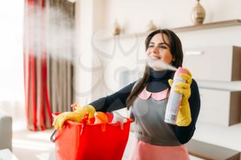 Smiling maid in uniform and rubber gloves  sprays air freshener in hotel room. Professional housekeeping equipment, charwoman