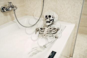 Scary human skeleton lying in the bathtub in foam, black humor, joke or surprise. Bathroom interior on background