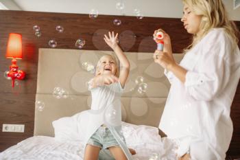 Funny mother and child blowing bubbles in bedroom at home. Parent feeling, togetherness, happy times
