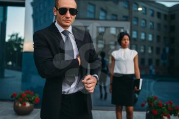 Serious bodyguard in suit, sunglasses and earpiece, female client on background. Security guard is a risky profession, professional guarding of business persons