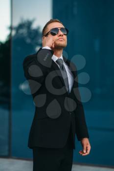 Male bodyguard talking by earpiece outdoors, security communication tools. Professional guarding is a risky profession. Guard in suit and sunglasses