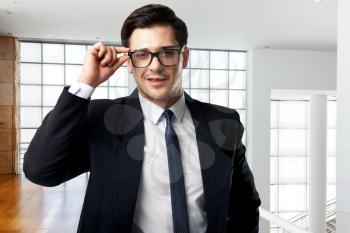 Young serious businessman in glasses, tie and black suit poses in modern business center with glass walls