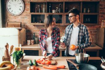 Young husband juggles oranges on the kitchen while wife cooking fruit breakfast. Man and woman preparing vegetable salad, happy family together