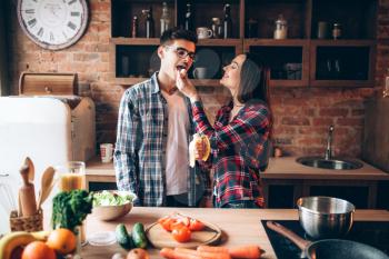 Young wife feeds husband a banana on the kitchen while cooking breakfast. Man and woman preparing vegetable salad, happy family together