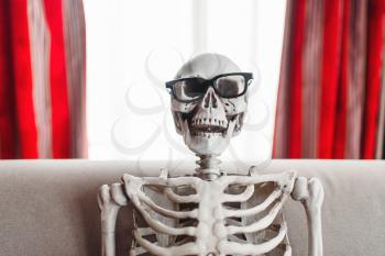 Smiling skeleton in glasses is sitting on couch, window and red curtains on background. Funny joke