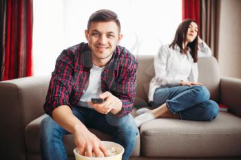 Couple sitting on the couch, man with remote control in hand watch tv and eats popcorn, woman in a bad mood, window on background