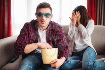 Couple sitting on the couch, man in 3D glasses with popcorn in hands watch movie, frightened woman closes her face with hands, window on background