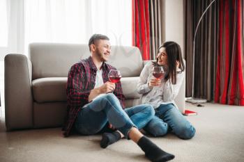 Love couple sitting on the floor against couch, watches movie and drinks red wine from big glasses, window and living room interior on background. Romantic evening