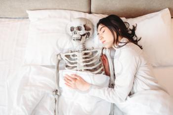 Young woman sleeping in bad with funny human skeleton together, top view. Humor or joke concept