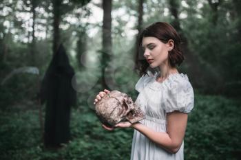 Female victim in white dress holds human skull in hands in the forest and death. Photo in horror style