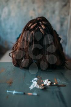Female junkie sitting at the table with drugs and syringe, grunge room interior on background. Drug addiction concept, addicted people