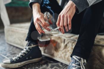Addicted male person sitting on the mattress with cigarette and bottle of alcohol in hands, grunge room interior on background. Addiction concept