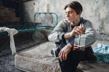 Male druggy sitting on the mattress, smoke cigarette and drinks alcohol, grunge room interior on background. Addiction concept