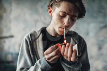 Male drug addict light a cigarette, grunge room interior on background. Smoking addiction concept