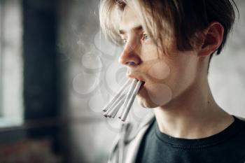 Young junkie with many cigarettes in his mouth reaching his hand forward, grunge background. No smoking concept, drug addict