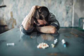 Male junkie sitting at the table with drugs and syringe, grunge room interior on background. Drug addiction concept, addicted people