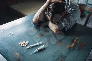 Male junkie sitting at the table with drugs and syringe, grunge room interior on background. Drug addiction concept, addicted people