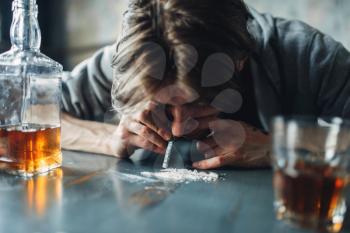 Male junkie sniffing a line of cocaine, grugs and bottle of alcohol on the table on background. Addiction concept, addicted people