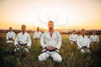 Karate group sitting on the ground and meditates, training in summer field. Martial art school on workout outdoor, relaxation in nature