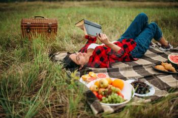 Cute woman lies on plaid and reads book, picnic on meadow. Romantic junket, happy holiday