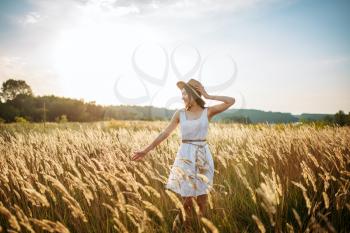 Cute woman in white dress and straw hat harvests wheat in the field. Pretty girl on summer meadow