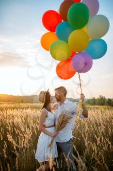 Happy couple holds bunch of balloons and kissing in wheat field. Pretty couple leisure on summer meadow