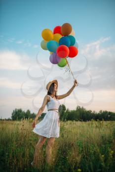 Smiling woman with colorful balloons walking in green field. Pretty girl on summer meadow at sunny day