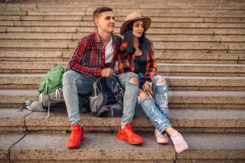 Couple of tourists with backpacks resting on the stone steps, excursion in town. Summer hiking. Hike adventure of young man and woman, city walking