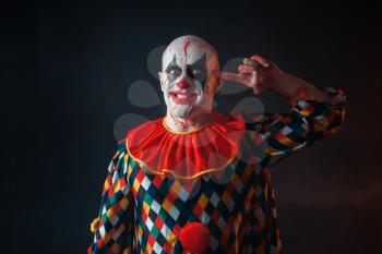 Portrait of mad bloody clown twirls finger to his temple, face in blood. Man with makeup in halloween costume, crazy maniac 
