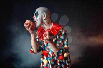 Mad bloody clown licks human brain. Man with makeup in carnival costume, crazy maniac holds organs in hands