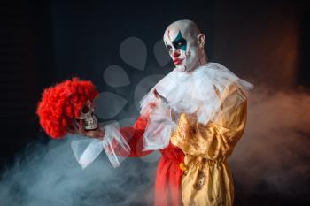 Mad bloody clown holds human skull in red wig, horror. Man with makeup in carnival costume, crazy maniac