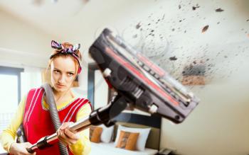 Angry housewife in apron with vacuum cleaner, flying pieces of dirt, bedroom interior on background. Crazy female person performs general cleaning, ruthless fight against dust