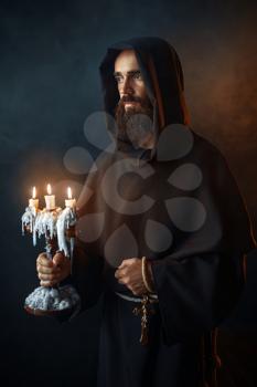 Medieval monk in robe holds a candlestick in hands, black background, secret ritual. Mysterious friar with candles. Mystery and spirituality