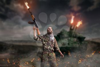 Bearded terrorist with rifle in hands stands in explosion and fire. Terrorism and terror, soldier in khaki camouflage