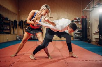 Woman makes elbow kick, self-defense workout with male personal trainer, gym interior on background. Female person on training, self defense practice
