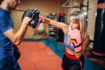 Female kickboxer on workout with male personal trainer in pads, gym interior on background. Woman boxer makes hand punch on training, kickboxing practice