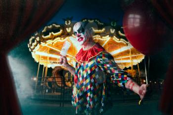 Mad bloody clown with meat cleaver holds air balloon, amusement park on background. Man with makeup in carnival costume, crazy maniac