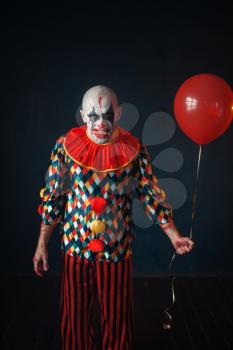 Ugly bloody clown with human finger in his teeth holds air balloon, horror. Man with makeup in carnival costume, crazy maniac