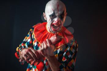 Mad bloody clown holds human hand, finger in his teeth. Man with makeup in halloween costume, crazy maniac