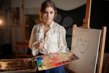 Female painter with color palette and brush standing against easel in studio. Creative paint, woman drawing pencil sketch, workshop interior on background