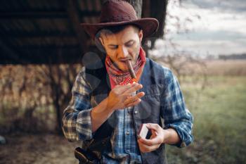 Brutal cowboy lights a cigar with matches, texas ranch on background, western. Vintage male person relax on farm, wild west lifestyle