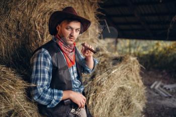 Brutal cowboy smokes a cigar, haystack on background, western. Vintage male person with gun on farm, wild west culture