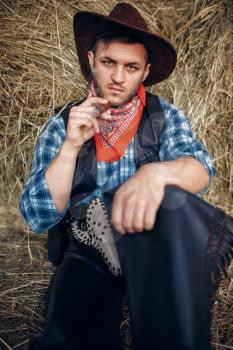 Brutal cowboy relax with cigar, haystack on background, western. Vintage male person with gun on farm, wild west culture