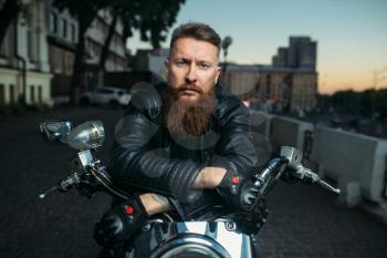 Brutal bearded biker poses on chopper in city, front view. Vintage bike, rider on motorcycle
