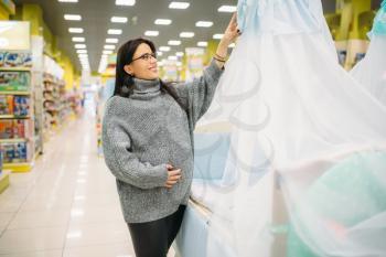 Pregnant woman choosing baby cot in store. Future mother in shop of goods for newburns