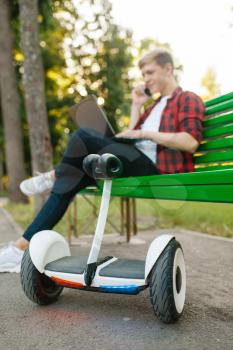 Young man with gyroboard sitting on the bench in park. Outdoor recreation with electric gyro board. Eco transport with balance technology, electrical gyroscope vehicle
