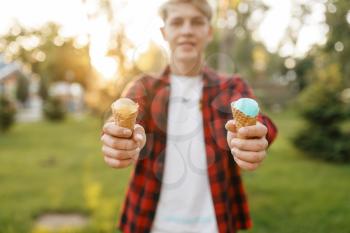 Young man holds out two ice creams in his hands in summer park. Youngster with ice-cream outdoors, male teenager with cold sweet